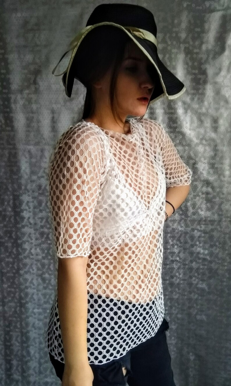 May include: A white mesh top with a round pattern. The top is sheer and has short sleeves. The model is wearing a white bra underneath the top.