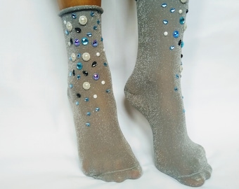 Può includere: Un paio di calzini grigio argento con finitura scintillante, adornati con una serie di abbellimenti decorativi. I calzini presentano una varietà di gemme, tra cui perle e cristalli blu, neri e trasparenti, aggiungendo un tocco di glamour.