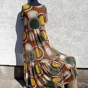 May include: A brown maxi dress with a repeating pattern of large circles in shades of brown, green, yellow, and white. The dress has short sleeves and a tiered skirt. The dress is displayed on a mannequin.