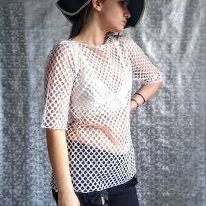 May include: A white mesh top with a round pattern. The top has short sleeves and a loose fit. The model is wearing the top with black jeans and a black hat.