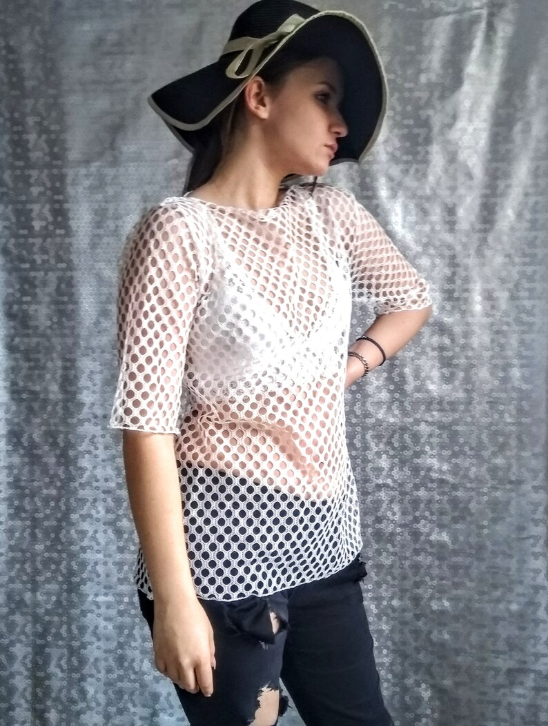 May include: A white mesh top with a round pattern. The top has short sleeves and a loose fit. The model is wearing the top with black jeans and a black hat.