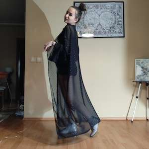 May include: A woman wearing a black sheer dress with polka dots and black boots. The dress has long sleeves and a flowing skirt. The woman is standing in a room with a map on the wall and a wooden easel with a painting on it.