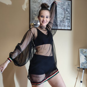 May include: A woman wearing a black sheer top with long sleeves and a black skirt. The top has a black bra underneath and a colorful stripe around the bottom. The woman is smiling and looking at the camera.