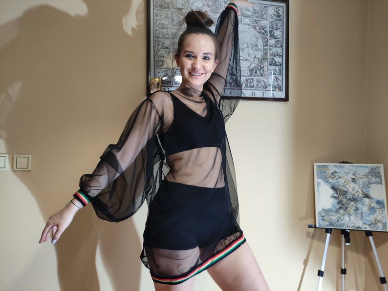 May include: A woman wearing a black sheer top with long sleeves and a black skirt. The top has a black bra underneath and a colorful stripe around the bottom. The woman is smiling and looking at the camera.