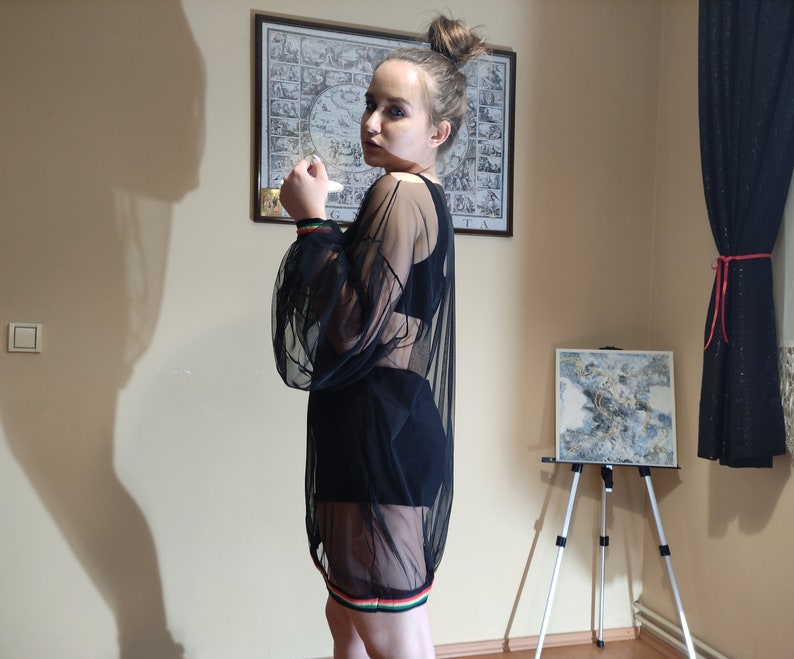 May include: A woman wearing a black sheer top with a black skirt and colorful trim. The top has a scoop neck and long sleeves. The skirt is a mini skirt with a high waist. The woman is standing in a room with a painting on the wall and a easel with a canvas on it.
