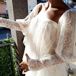 Peut inclure: Une robe blanche à manches transparentes brodées de motifs floraux. La robe présente une encolure carrée et un design fluide à plusieurs niveaux. Les manches sont longues et bouffantes, avec des motifs floraux complexes. La robe semble être faite d'un tissu délicat et léger.