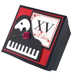 Puede incluir: Una caja decorativa negra y roja con un diseño de piano y máscara. La caja presenta un teclado de piano, una máscara y una pequeña rosa roja. La caja también tiene el texto "XV Anos" en rojo.