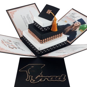 以下が含まれることがあります： 卒業をテーマにしたポップアップカード。白と黒を基調としたデザインで、装飾的な箱の積み重ね、卒業帽子、卒業生の写真が特徴です。「Grad」の文字は金色のグリッターで表示されています。