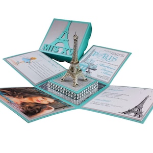 Può includere: Una scatola esplosiva turchese e argento con un centrotavola a forma di Torre Eiffel dorata. La scatola presenta inviti stampati con il testo "Paris" e "Mis XV". La scatola è decorata con strass e una foto.