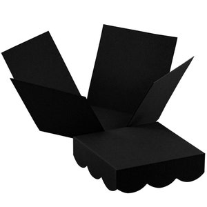 Può includere: Una scatola esplosiva nera, aperta e visualizzata. La scatola è quadrata con bordi smerlati sul fondo. I lati della scatola sono ripiegati, rivelando l'interno. La scatola è realizzata in un materiale nero opaco.