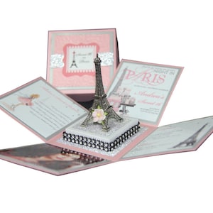 Può includere: Una scatola di invito rosa e argento per Sweet 16 con un centrotavola a forma di Torre Eiffel. La scatola si apre per rivelare biglietti d'invito con testo e illustrazioni. La Torre Eiffel è argentata con un fiore rosa.