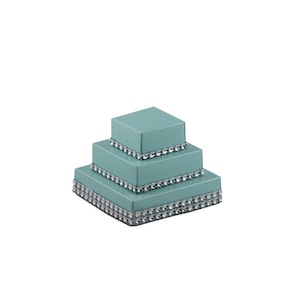 Può includere: Un set di scatole regalo quadrate a tre livelli, di colore turchese. Ogni scatola è decorata con un bordo di strass argentati. Le scatole sono impilate, creando un effetto a più livelli, ideale per occasioni speciali.