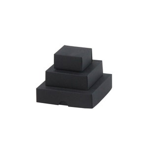 Può includere: Tre scatole regalo quadrate nere di diverse dimensioni, impilate l'una sull'altra. Le scatole hanno una finitura strutturata e un design minimalista. La scatola più piccola è in cima, seguita da una scatola di medie dimensioni e da una scatola più grande in basso.