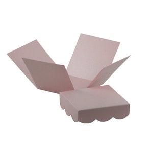 Può includere: Una scatola regalo quadrata rosa chiaro, aperta, con bordi smerlati. I lati della scatola sono aperti, rivelando l'interno. La scatola è fatta di carta o cartoncino e si trova su uno sfondo bianco.