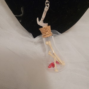 May include: A silver necklace with a small glass bottle pendant containing red and white beads and two small white bones. The bottle is topped with a cork and a silver hammer charm hangs from the top of the cork.