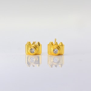 Puede incluir: Un par de pendientes dorados con forma de cámaras en miniatura. Cada cámara tiene una gema redonda y transparente en el centro de la lente. Los pendientes se muestran sobre una superficie reflectante.