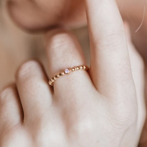 May include: A delicate gold ring with a single small pink gemstone set in the center. The ring is made of tiny gold beads.