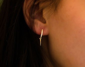May include: Gold hoop earrings with a small, dangling spike charm. The earrings are set with clear cubic zirconia stones.