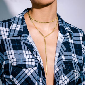 May include: A gold necklace with two strands, a choker and a long, Y-shaped pendant. The necklace is worn over a blue and black plaid flannel shirt.