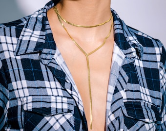 May include: A gold-colored necklace with two strands, one a choker and the other a long, Y-shaped pendant. The necklace is worn over a blue and black plaid flannel shirt.