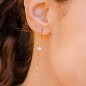May include: A gold-colored earring with a delicate chain design. The earring features a small, clear gemstone and a straight bar at the end. The earring is shown worn on an ear.