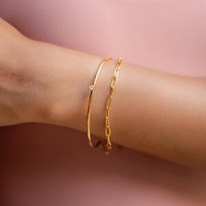 May include: Two gold bracelets on a person's arm. One is a delicate bangle with a small, clear gemstone. The other is a chain link style bracelet. The arm is against a pink background.