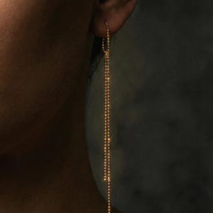 May include: Gold-colored, long dangle earrings. The earrings are made of small, round beads strung together to create a delicate, flowing design. The earrings are shown against a dark background.