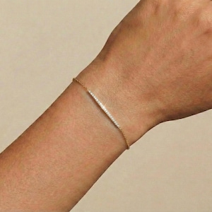 May include: A gold bracelet with a bar of small, clear stones. The bracelet is on a wrist with a neutral background.