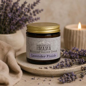 May include: A jar of "Mountain Mama Whipped Tallow Cream - Lavender Fields" with a gold lid sits on a ceramic plate, surrounded by lavender sprigs. A lit candle and a vase of lavender are in the background.