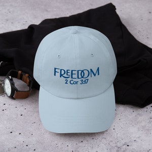 May include: A pale blue baseball cap featuring the word "FREEDOM" embroidered in dark blue, with the text "2 Cor 3:17". The cap is made of a soft material and has a curved peak.