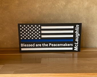 May include: Black and white American flag with a blue stripe across the middle. The text "Blessed are the Peacemakers" is written below the flag. The name "McLaughlin" is written vertically on the right side of the flag.