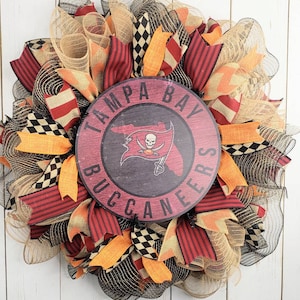 May include: A wreath decorated with black, orange, and red ribbon, burlap, and checkered fabric. The wreath features a wooden sign with the text "Tampa Bay Buccaneers".