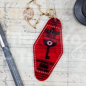May include: Red motel key chain with a black and white illustration of a key with a heart-shaped eye and wings. The text "Hazbin Hotel" and "Pentagram City" are printed on the key chain.