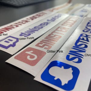 May include: A collection of four rectangular adhesive decals featuring the text SINISTER SOCIETY and various social media icons. The decals are available in glitter red, glitter purple, glitter pink, and a reflective chrome blue finish.