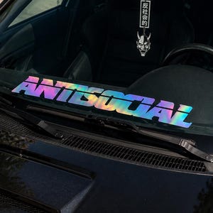 May include: A holographic decal applied to the lower windshield of a black car, displaying the word ANTISOCIAL in bold, block capital letters. A hanging rearview mirror ornament features a white oni mask and vertical Japanese text characters.