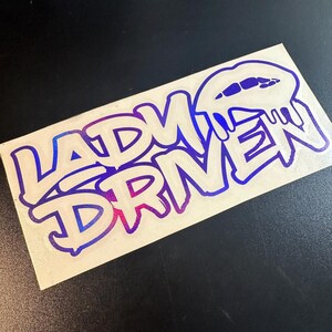 Pu&ograve; includere: Un adesivo bianco con un design sfumato blu e rosa. L'adesivo presenta il testo "Lady Driven" in un font in stile graffiti. La "D" di "Driven" ha la forma di una labbra che cola.