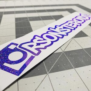 May include: A rectangular white sticker featuring the text "RSONSTEROIDS" in a bold, stylized purple holographic font. The lettering has a glittery, reflective finish that shifts in color, set against a plain white background on the sticker.