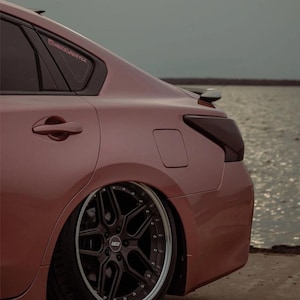 May include: A rose gold sedan featuring a lowered suspension, dark multi-spoke alloy wheels with a polished lip, and tinted taillights. A small sticker is visible on the rear passenger window, and the car is positioned near a body of water.