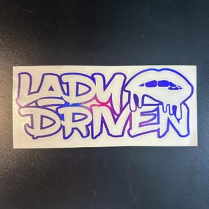 Pu&ograve; includere: Un adesivo bianco con le parole "Lady Driven" in un gradiente blu e viola. Le parole sono in un font in stile graffiti. Un labbro blu e viola con gocce &egrave; a destra delle parole.