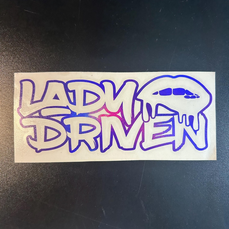 Pu&ograve; includere: Un adesivo bianco con le parole "Lady Driven" in un gradiente blu e viola. Le parole sono in un font in stile graffiti. Un labbro blu e viola con gocce &egrave; a destra delle parole.