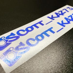 May include: A rectangular white sticker featuring a blue gradient Instagram camera icon followed by the text SCOTT_KAZ73 in a bold, sans-serif font. The design is repeated twice on the sticker, with the text and icon showing a metallic blue finish.