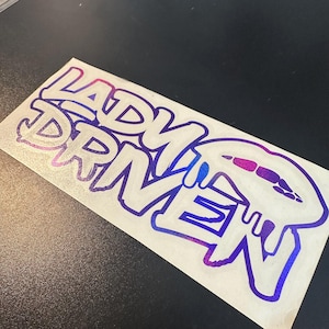 Pu&ograve; includere: Un adesivo con le parole "Lady Driven" in un font di stile graffiti. L'adesivo &egrave; un gradiente di viola e blu.