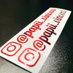 May include: Two rectangular white vinyl decals featuring the red metallic Instagram logo icon followed by the text @papii_vincent in a cursive script font. The decals are arranged diagonally on a dark surface, displaying a glossy finish.