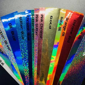Pu&ograve; includere: Una variet&agrave; di fogli di vinile iridescente con diverse finiture, tra cui glitter, effetto olio, cromo e metallo spazzolato. I fogli sono etichettati con i loro nomi, come "Glitter Black", "Black Oil", "Chrome Red", "Orange Oil", "Gold Oil", "Gold", "Pink Chrome", "Pink Glitter", "Glitter Green", "Chrome Blue", "Glitter Blue", "Oil Blue", "Glitter Silver", "Oil Slick" e "Brush Metal".