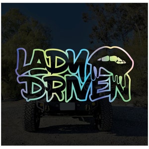Pu&ograve; includere: Una Jeep nera con un adesivo colorato "Lady Driven" sul lunotto posteriore. L'adesivo presenta un design di rossetto che cola.