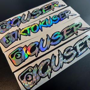 May include: Several holographic stickers with the word "GIGUSER" in a stylized font. The stickers have a reflective, rainbow-like appearance. The word "TIKTOKUSER" is also visible on one sticker. The stickers are on a white background.