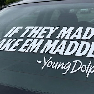 May include: A white vinyl decal applied to a car rear window featuring the text IF THEY MAD MAKE EM MADDER in a bold sans-serif font, followed by the name -Young Dolph in a cursive script font, creating a high-contrast graphic display.