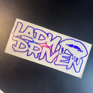 Pu&ograve; includere: Un adesivo bianco con le parole "Lady Driven" in un font degradato blu e viola. Le parole sono in un font in stile graffiti. L'adesivo ha un design a labbra gocciolanti.