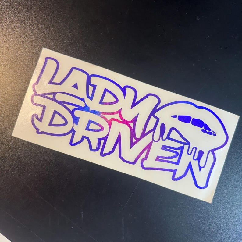 Pu&ograve; includere: Un adesivo bianco con le parole "Lady Driven" in un font degradato blu e viola. Le parole sono in un font in stile graffiti. L'adesivo ha un design a labbra gocciolanti.