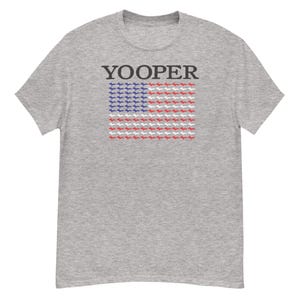 Peut inclure: T-shirt gris clair avec le mot "YOOPER" imprimé en haut. En dessous, un motif de drapeau composé de silhouettes bleues, blanches et rouges de la péninsule supérieure du Michigan, ressemblant au drapeau américain.